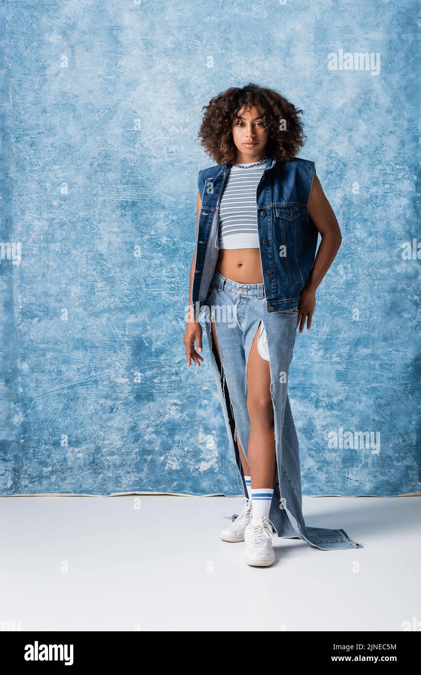 intera lunghezza di donna afroamericana alla moda in jeans strappati e giubbotto denim in piedi su sfondo blu Foto Stock