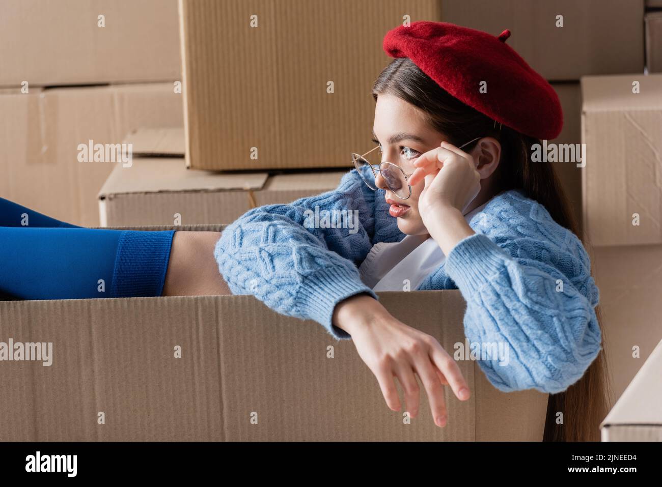 Modello piuttosto giovane in occhiali e berretto seduto in scatola di cartone Foto Stock