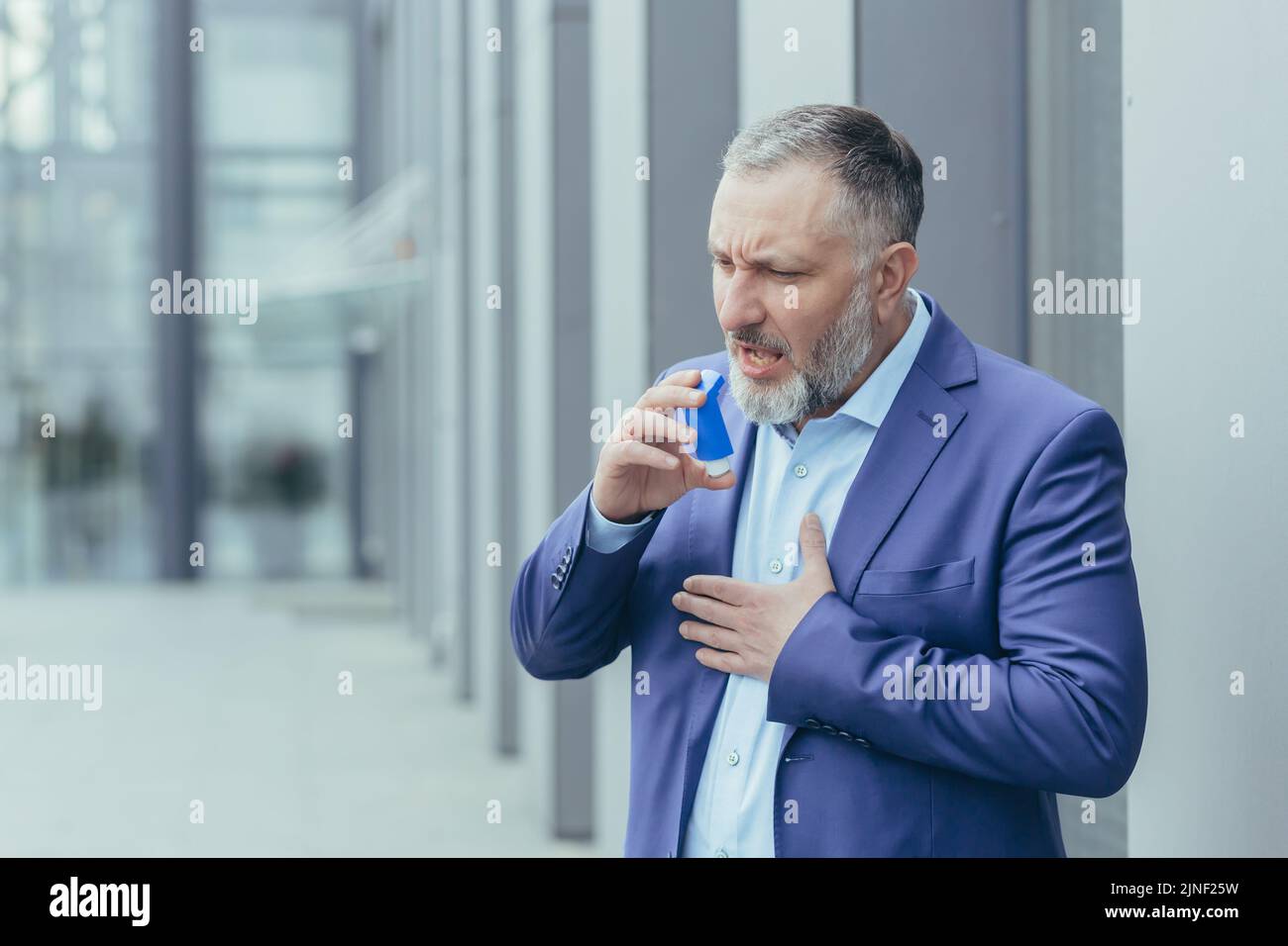 Uomo d'affari anziano grigio-haired malato ha attacco di asma difficile da respirare, uomo fuori ufficio edificio in vestiti di affari utilizza inalatore per facilitare la respirazione Foto Stock