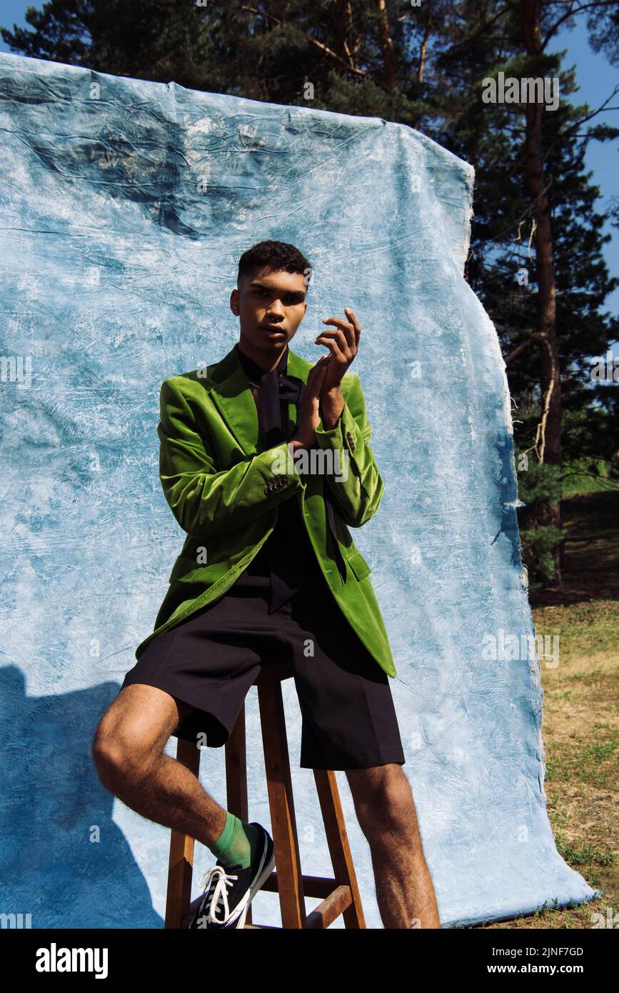 uomo alla moda afroamericano in blazer verde e pantaloncini che guardano la macchina fotografica mentre si siede vicino a stoffa blu all'aperto, immagine stock Foto Stock