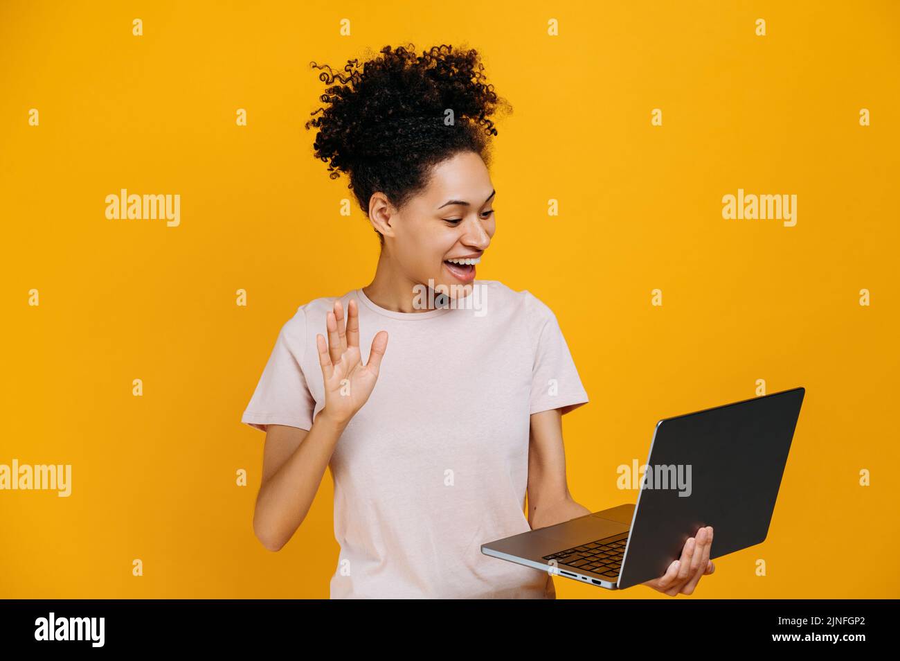 Comunicazione video online. Amichevole afroamericana donna, tenendo in mano un computer portatile aperto, facendo un gesto di saluto, parlando in videoconferenza, in piedi su sfondo arancione isolato, sorridendo Foto Stock