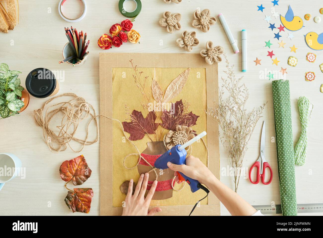 Direttamente sopra la vista di una donna irriconoscibile che utilizza la pistola colla a caldo mentre decorare l'elemento di decorazione con spago, la superficie della scrivania disordinata Foto Stock