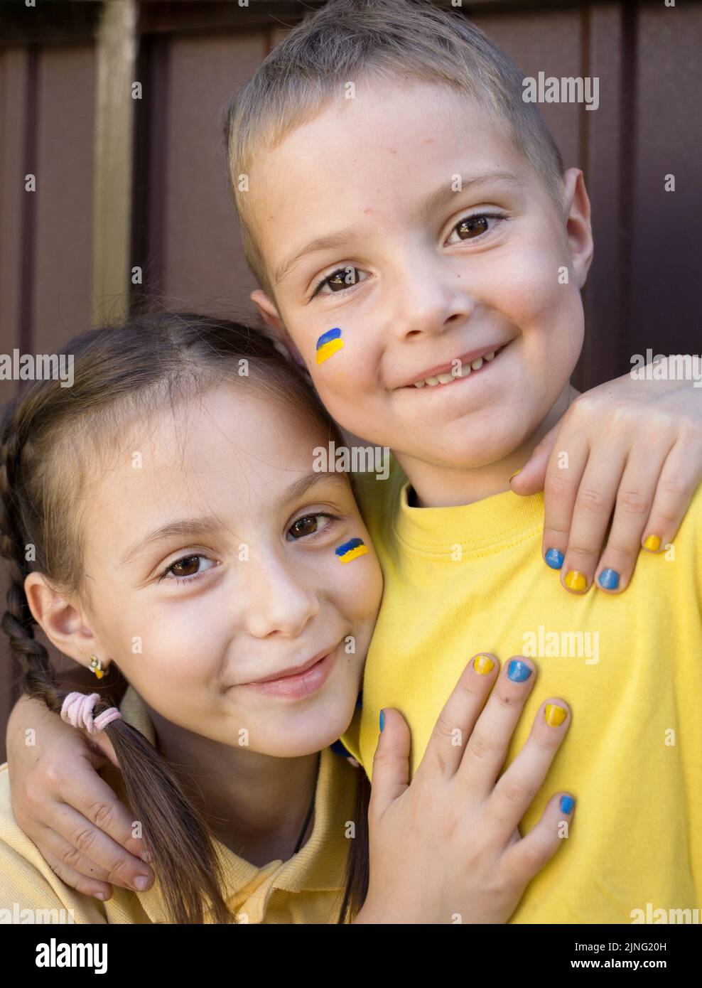 Facce di un felice ragazzo di 3 anni e di una ragazza di 7 anni in magliette gialle con bandiere dipinte di Ucraina sulle guance. Fratello e sorella abbracciare. Regno Unito Foto Stock