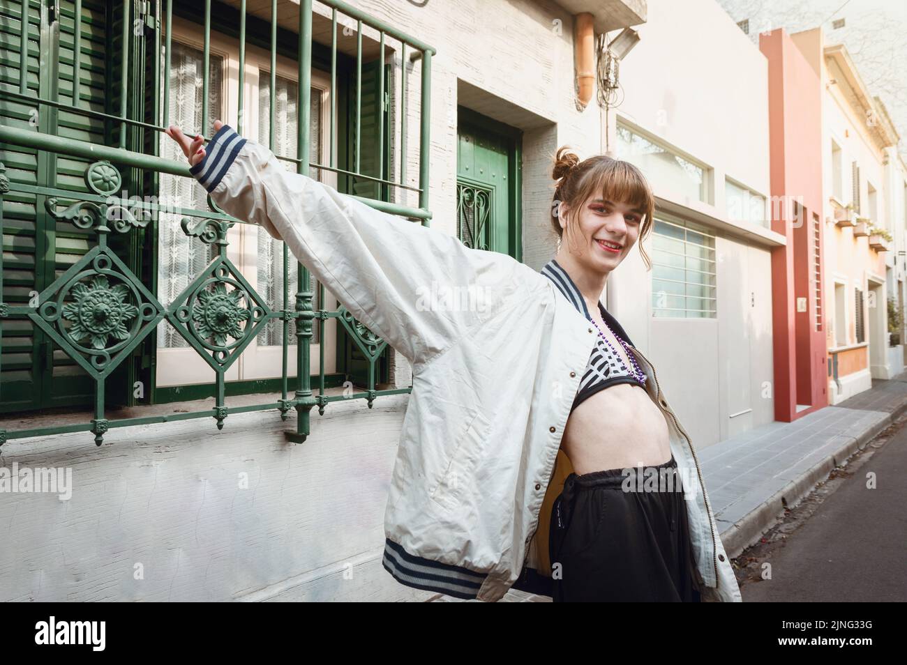felice giovane donna transgender latina con braccia aperte in piedi lateralmente sulla strada guardando la fotocamera sorridente, in un ambiente urbano con case Foto Stock