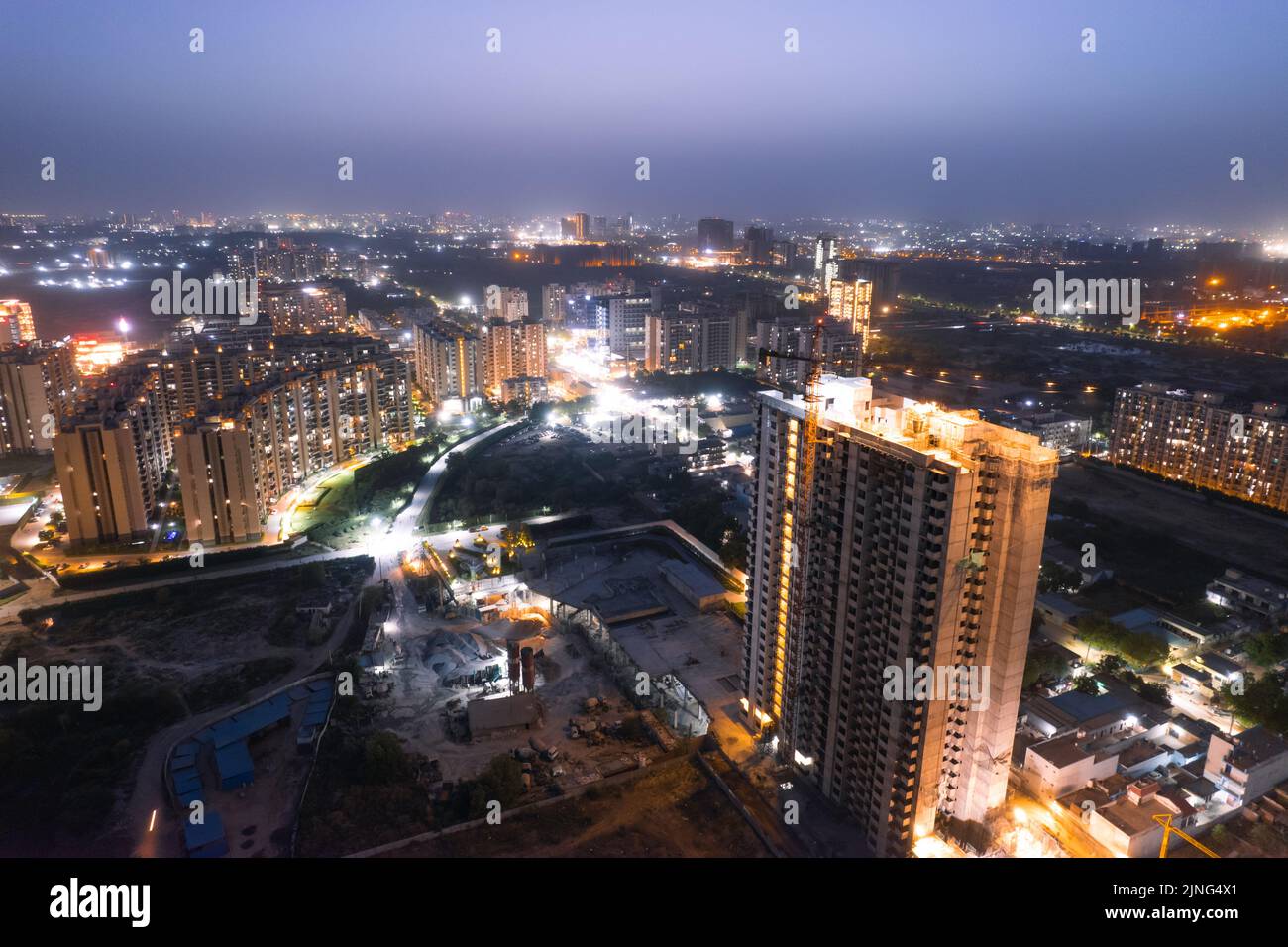 crepuscolo con droni aerei che mostra luci arancioni di strade, case e mercati che circondano un grattacielo con il paesaggio della città che si estende nel Foto Stock