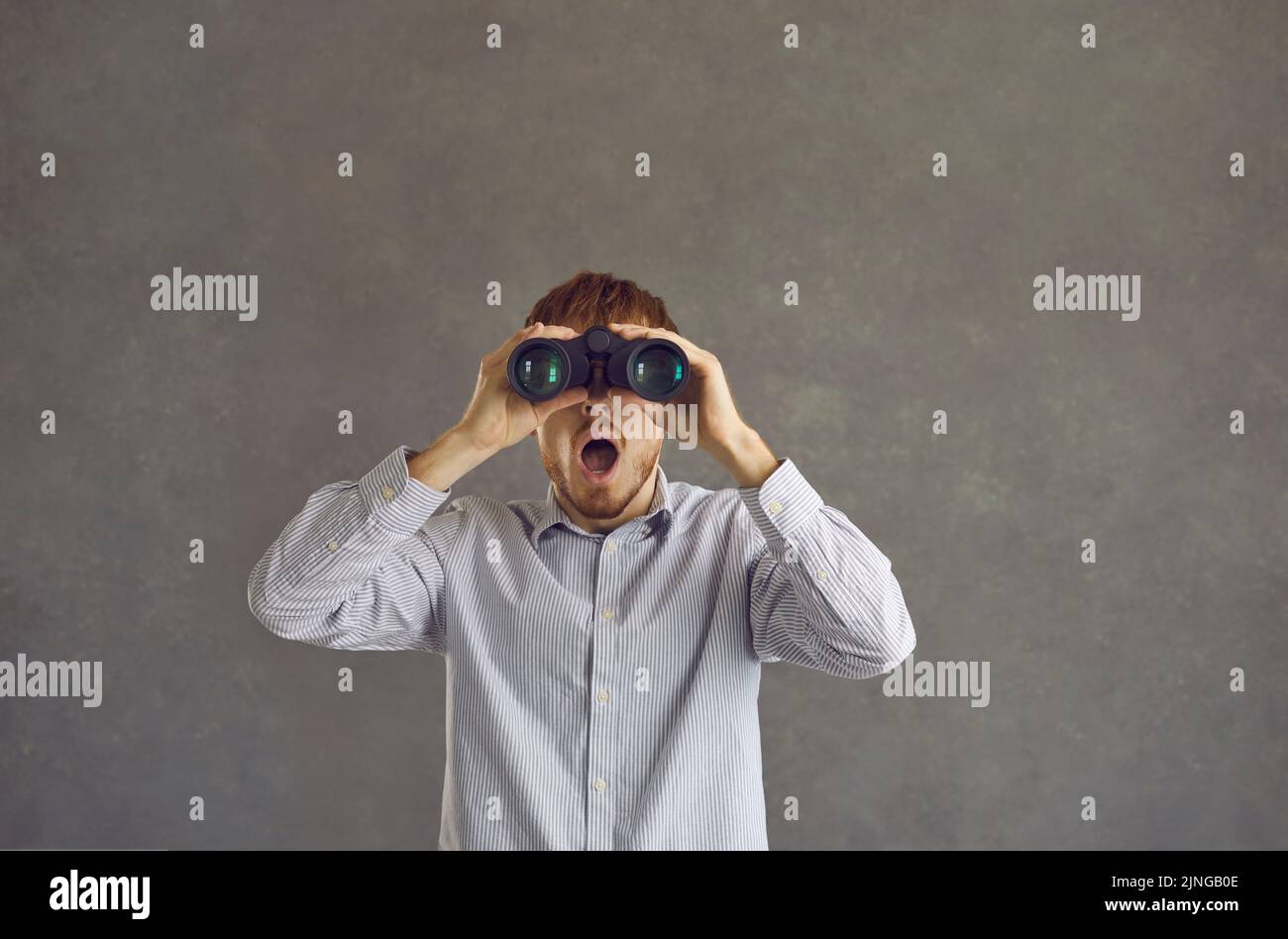 Uomo divertente che guarda a bocca aperta in binocolo in piedi isolato su sfondo grigio Foto Stock