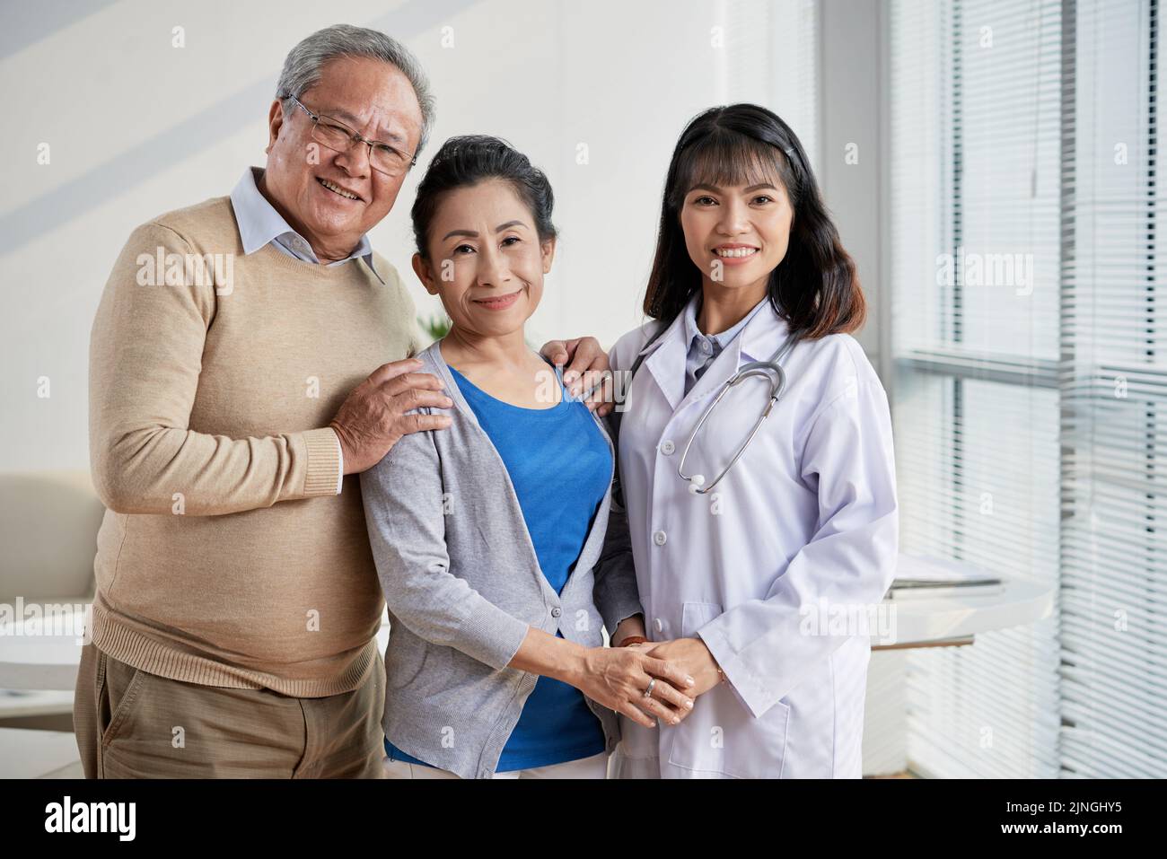 Ritratto di gruppo di un medico asiatico altamente professionale e di una coppia anziana affettuosa in piedi presso un ufficio moderno e guardando la macchina fotografica con sorrisi ampi Foto Stock