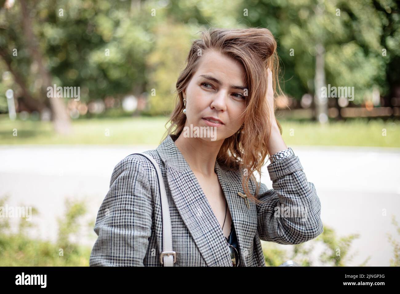 Primo piano ritratto di una giovane donna d'affari che tocca i capelli e guarda via nelle giornate di sole nel parco cittadino. Camminare per strada, stile di vita moderno, meditativo Foto Stock