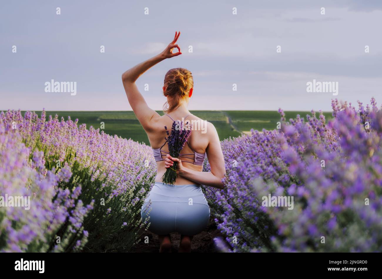 Donna in abbigliamento attivo che tiene un bouquet di lavanda in una posa yoga, in un campo di lavanda al tramonto con il segno ok sopra la testa Foto Stock