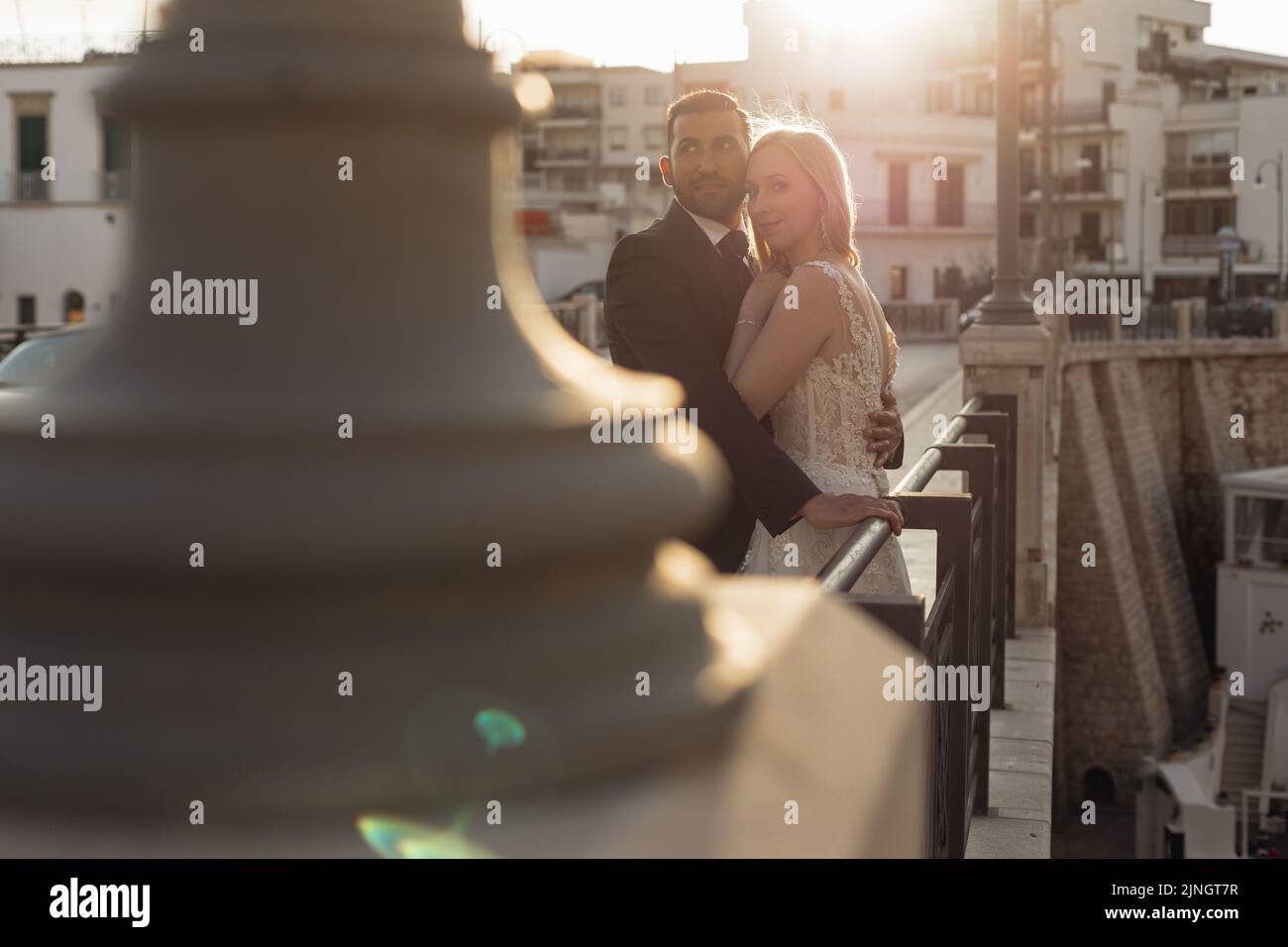 Vista laterale di una coppia di sposi in piedi sulla ringhiera di acciaio del ponte al tramonto. Giovane sposa abbracciante con lo sposo. Foto Stock