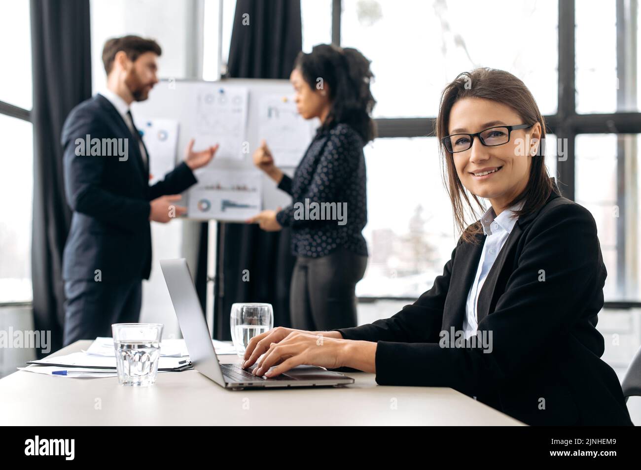 Sicura ed elegante donna di affari caucasica con occhiali, seduto alla scrivania alla conferenza, vicino al computer portatile. Bella donna elegante in riunione briefing, guardando la fotocamera, sorridente amichevole Foto Stock