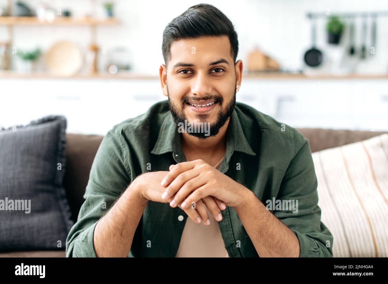 Ritratto di sicuro attraente indiano o arabo uomo, in abiti casual elegante, seduto a casa in accogliente soggiorno sullo sfondo della cucina, guarda la macchina fotografica, sorridendo amichevole Foto Stock