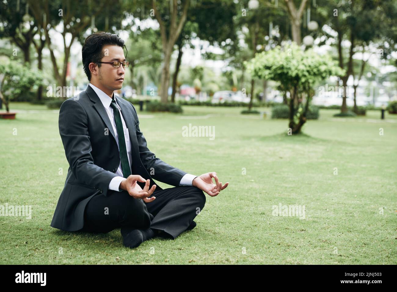 Giovane uomo d'affari asiatico in tuta praticando meditazione all'aperto nel parco Foto Stock