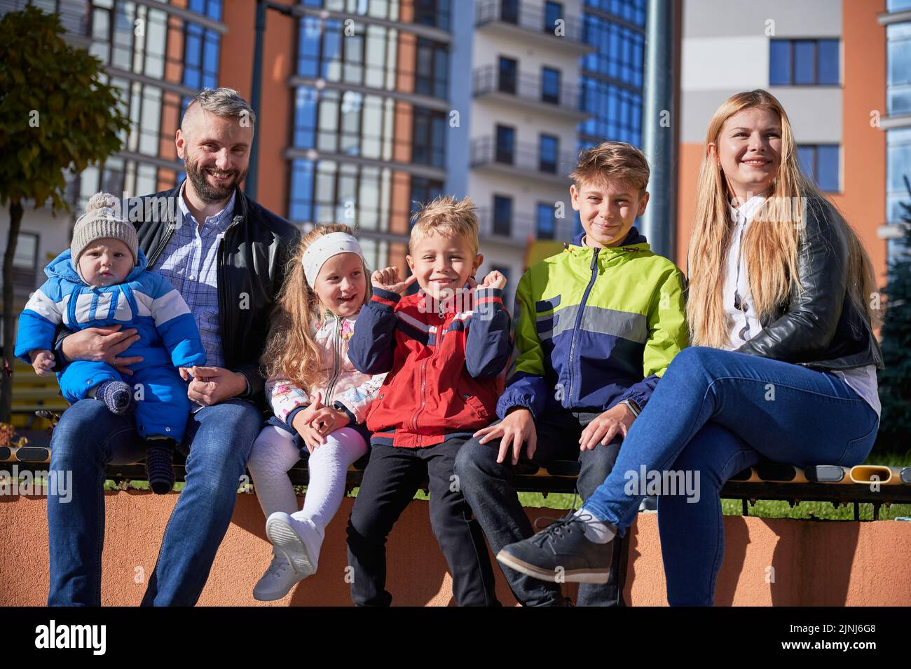 Due adulti che trascorrono del tempo con bambini carini, sorridendo nella giornata di sole primaverile. Vista frontale dei genitori di mezza età seduti sulla panchina con quattro adorabili bambini, riposati nel centro della città. Concetto di famiglia. Foto Stock