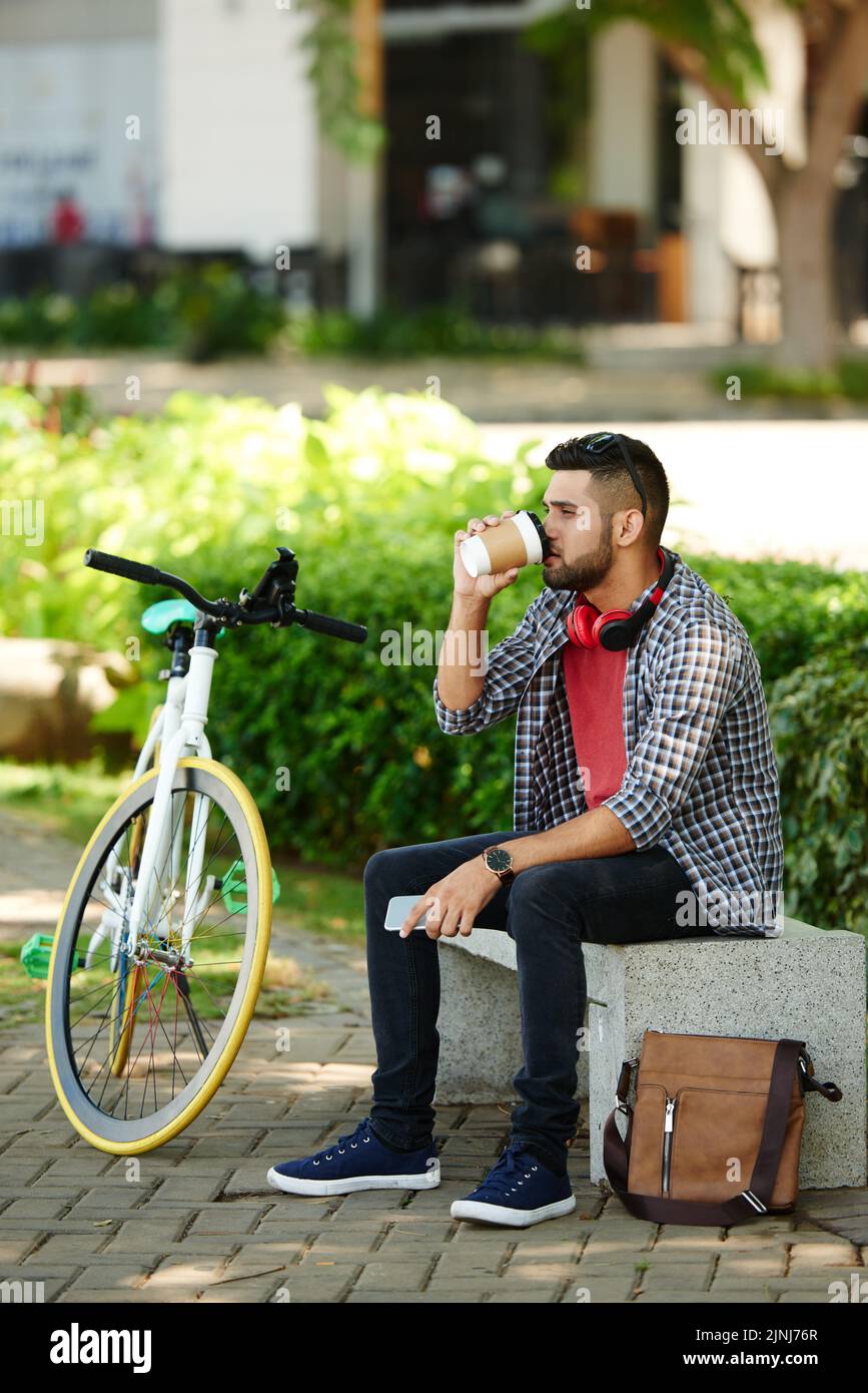Ciclista fiduciosa seduta sul panca del parco e gustando un caffè fragrante mentre si prende una breve pausa, ritratto completo Foto Stock