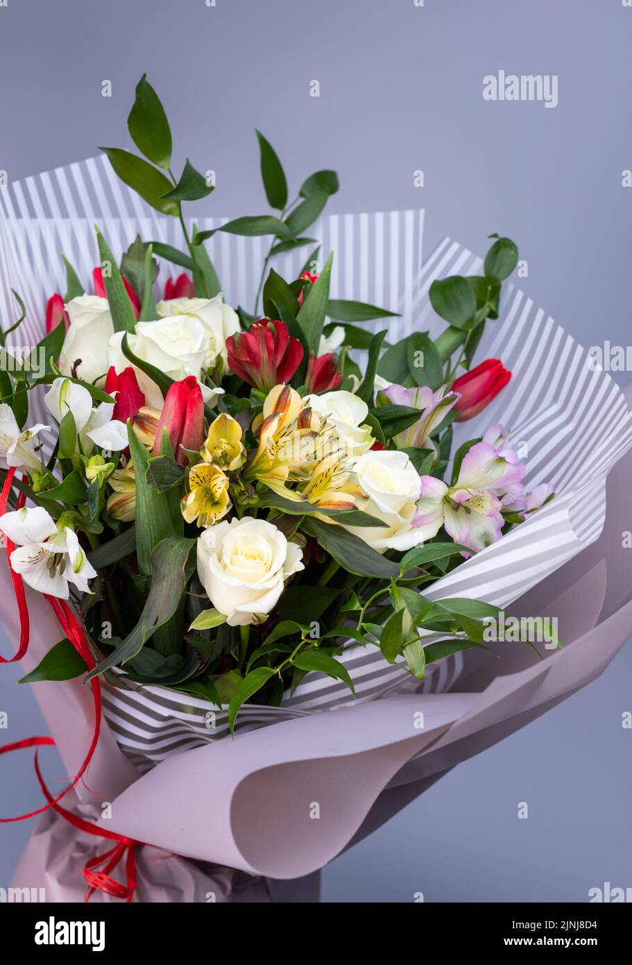 Primo piano di un luminoso bouquet di rose bianche, iridi colorati e tulipani rossi. Bouquet di fiori avvolto in carta floreale leggera di due tipi su un dorso blu Foto Stock