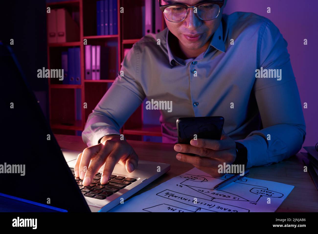 Un bel programmatore asiatico che indossa camicia e occhiali mettendo un tocco finale in una nuovissima app per computer mentre si è seduti alla scrivania in un ufficio poco luminoso, in un ritratto in vita Foto Stock