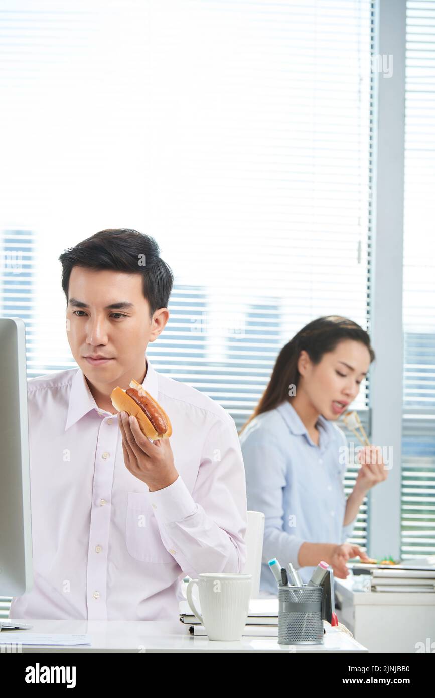 Giovane imprenditore vietnamita mangiare hot dog e lavorare sul computer in ufficio Foto Stock