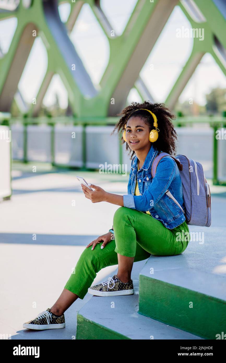 Ragazza adolescente multirazziale con zaino, cuffie e smartphone che riposa dopo la scuola nel ponte della città, ascoltando musica. Foto Stock