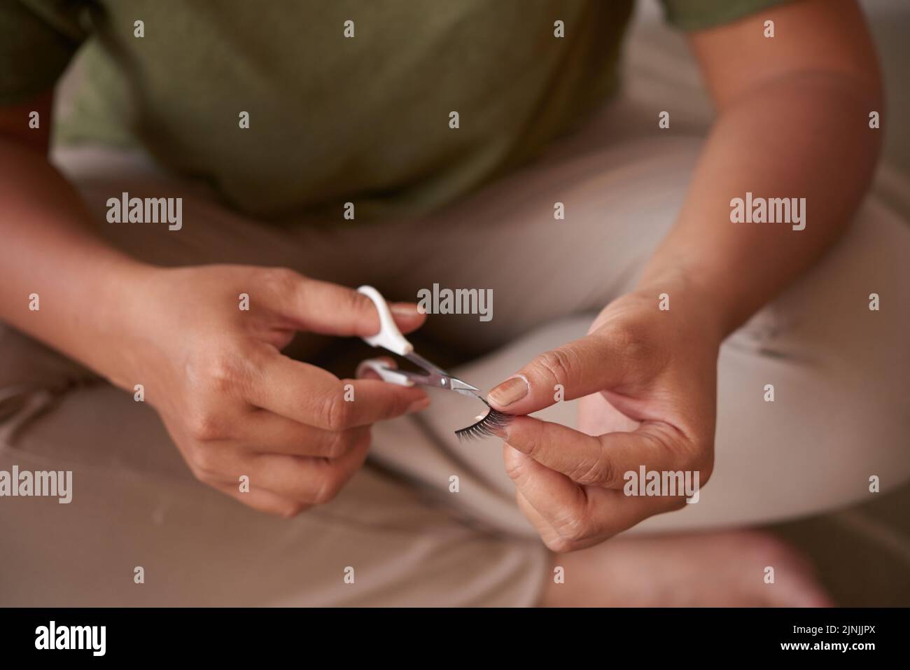 Artista di make-up irriconoscibile seduto in posizione lotus mentre si tagliano le lunghezze delle false ciglia con l'aiuto di forbici, primo piano Foto Stock