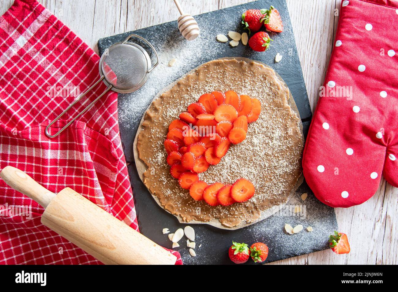 Torta di torta di galette fatta in casa con fragole e mandorle, posta su scrivania nera e tavolo di legno. Asciugamano rosso con utensili da cucina sul lato Foto Stock