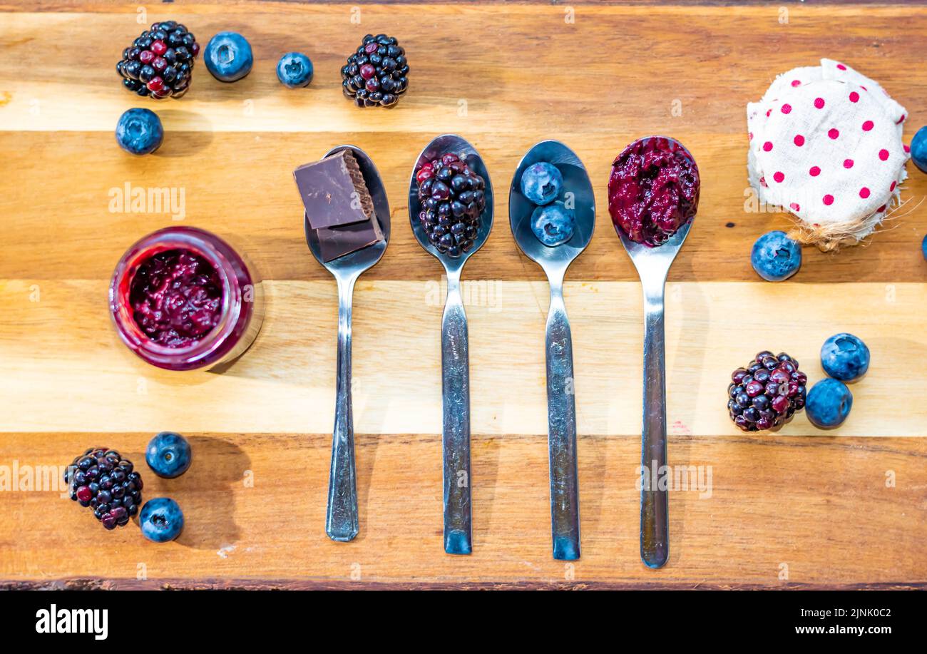 Mirtillo, mora e cioccolato sono messi in cucchiaio d'argento su scrivania di legno. Ingredienti per la marmellata. Frutta fresca intorno. Foto Stock