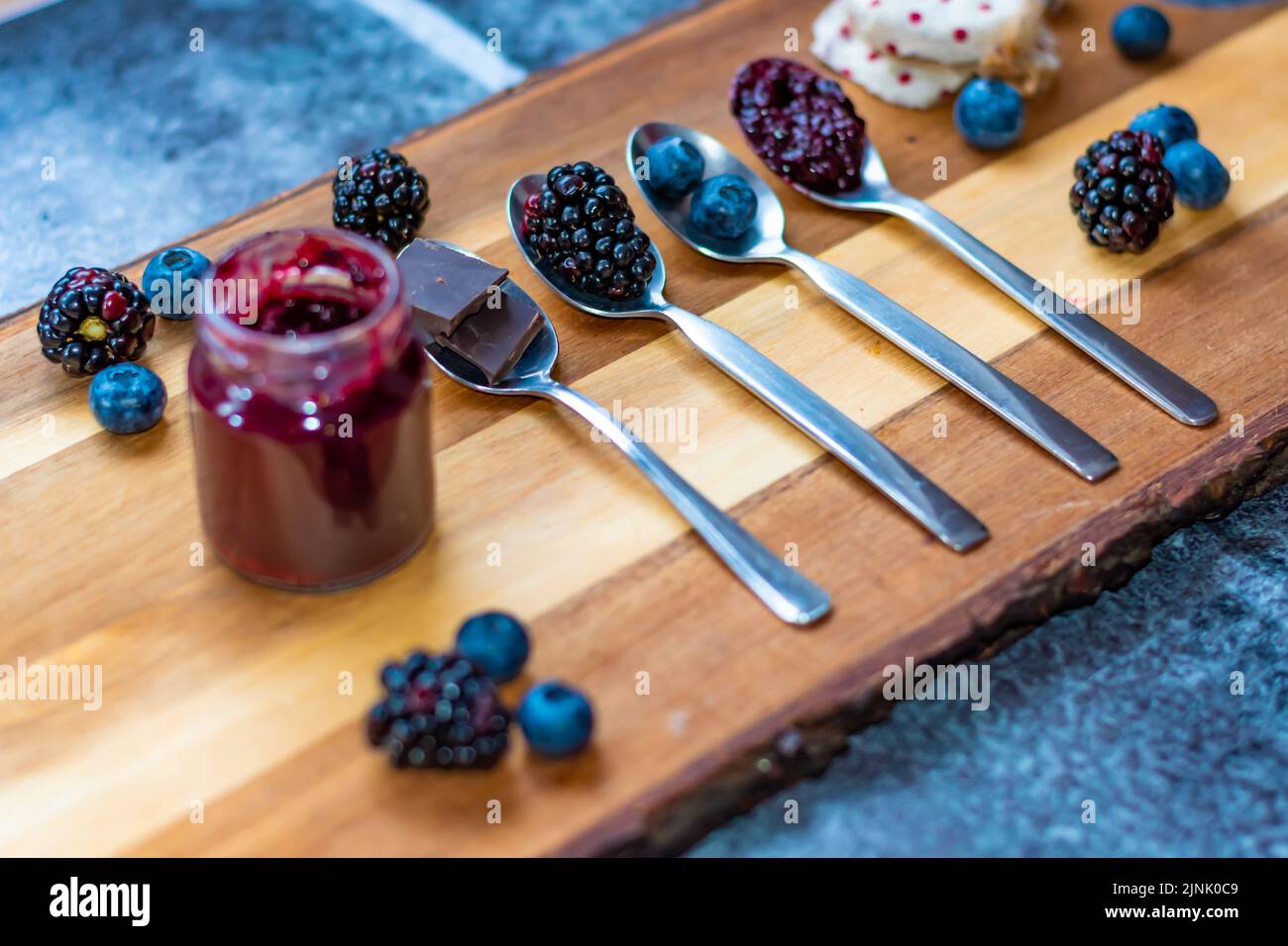 Mirtillo, mora e cioccolato sono messi in cucchiaio d'argento su scrivania di legno. Ingredienti per la marmellata. Frutta fresca intorno. Foto Stock