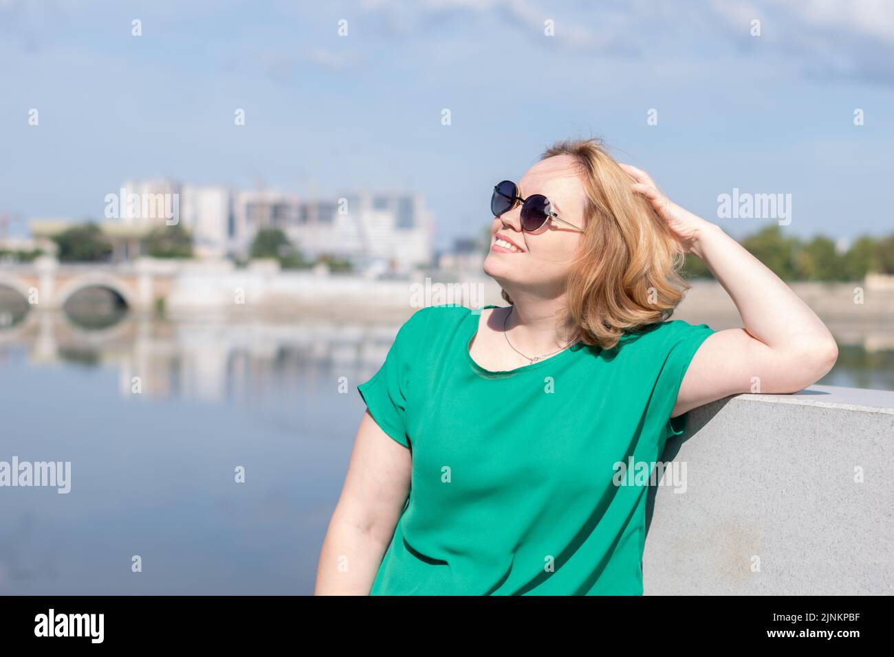Ritratto di una ragazza in occhiali da sole, una blusa verde in piedi sulla riva del fiume tenendo i capelli con la mano. Una ragazza cammina per le strade del cit Foto Stock