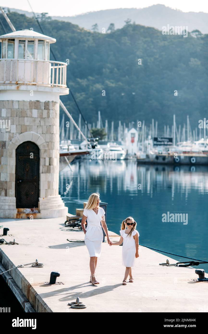 Felice madre millenaria e figlia bionda adolescente in abiti bianchi a piedi vicino al porto e porto turistico con yacht in una località turistica di mare con vela Foto Stock