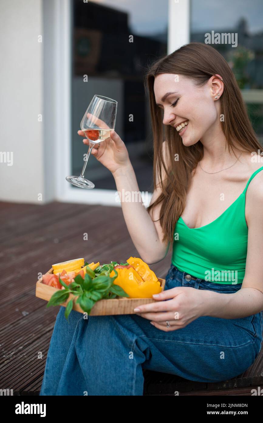 Ritratto di giovane donna sorridente beauteous seduta sul gradino di veranda di legno, tenendo un bicchiere di vino bianco tagliato verdure. Foto Stock