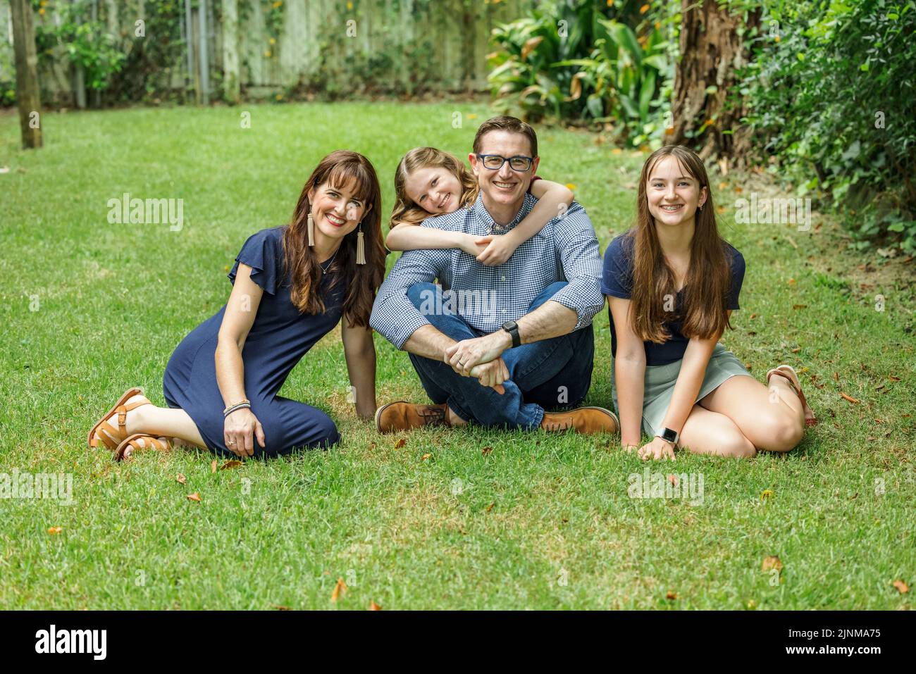 Una famiglia di una madre e di un padre e due figlie seduti all'esterno nell'erba del loro cortile, con la figlia più giovane che abbraccia il papà da dietro Foto Stock