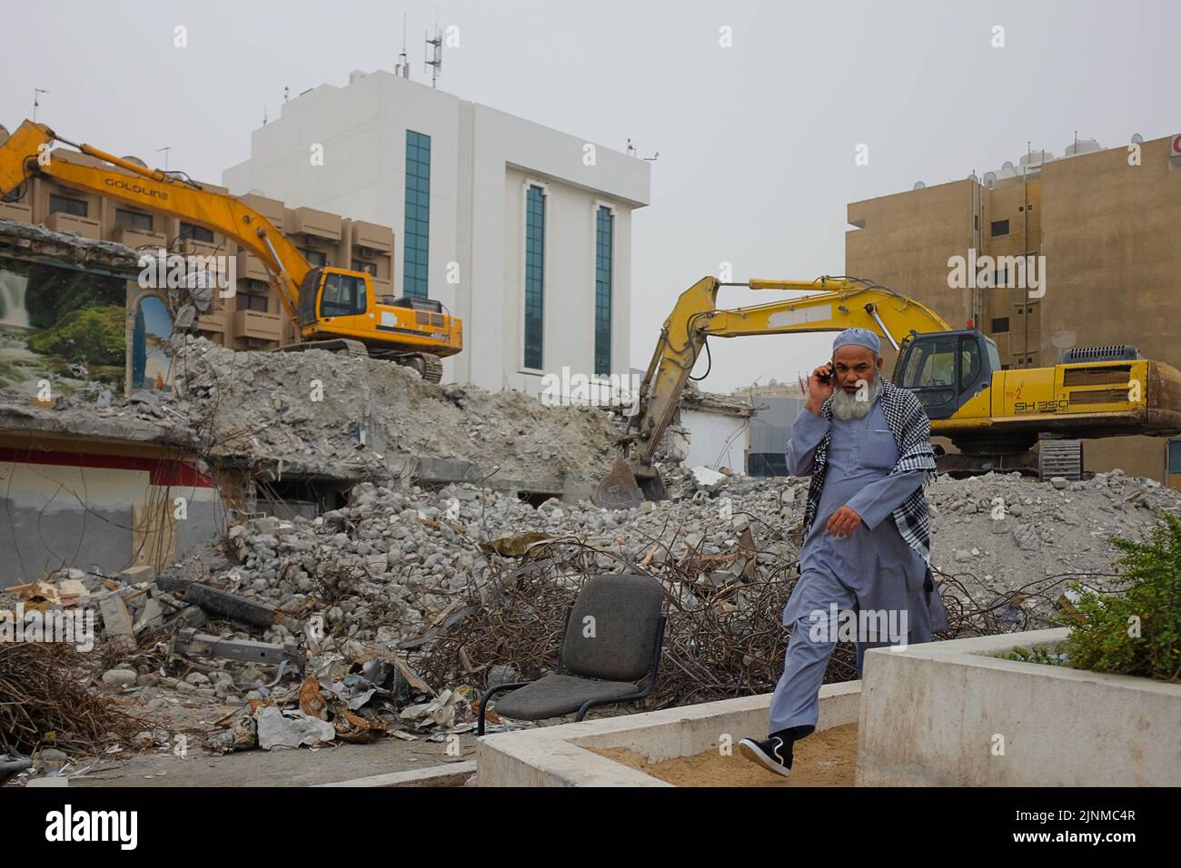 Un uomo pakistano su una chiamata cammina davanti al luogo di demolizione con due escavatori gialli ad al Ghubaiba. Scena diurna all'aperto a Dubai, Emirati Arabi Uniti. Foto Stock