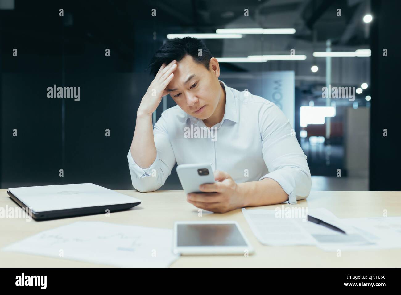 Un ragazzo asiatico confuso che tiene in mano uno smartphone si sente preoccupato di pensare al messaggio ricevuto. Utilizzo quotidiano del telefono cellulare, notizie spiacevoli, attesa di una chiamata importante, concetto di problemi di dispositivo con segnale basso Foto Stock