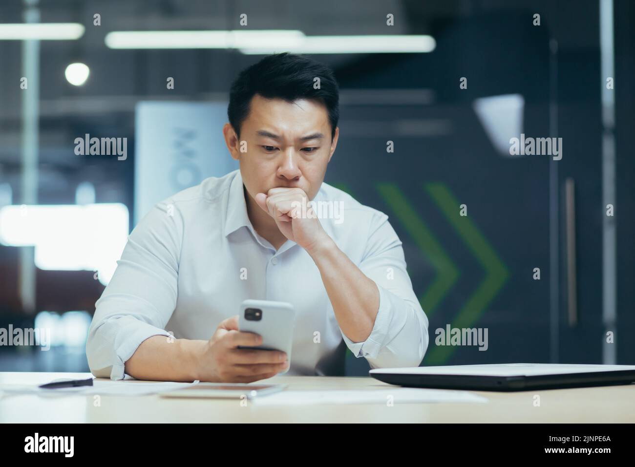 Un ragazzo asiatico confuso che tiene in mano uno smartphone si sente preoccupato di pensare al messaggio ricevuto. Utilizzo quotidiano del telefono cellulare, notizie spiacevoli, attesa di una chiamata importante, concetto di problemi di dispositivo con segnale basso Foto Stock