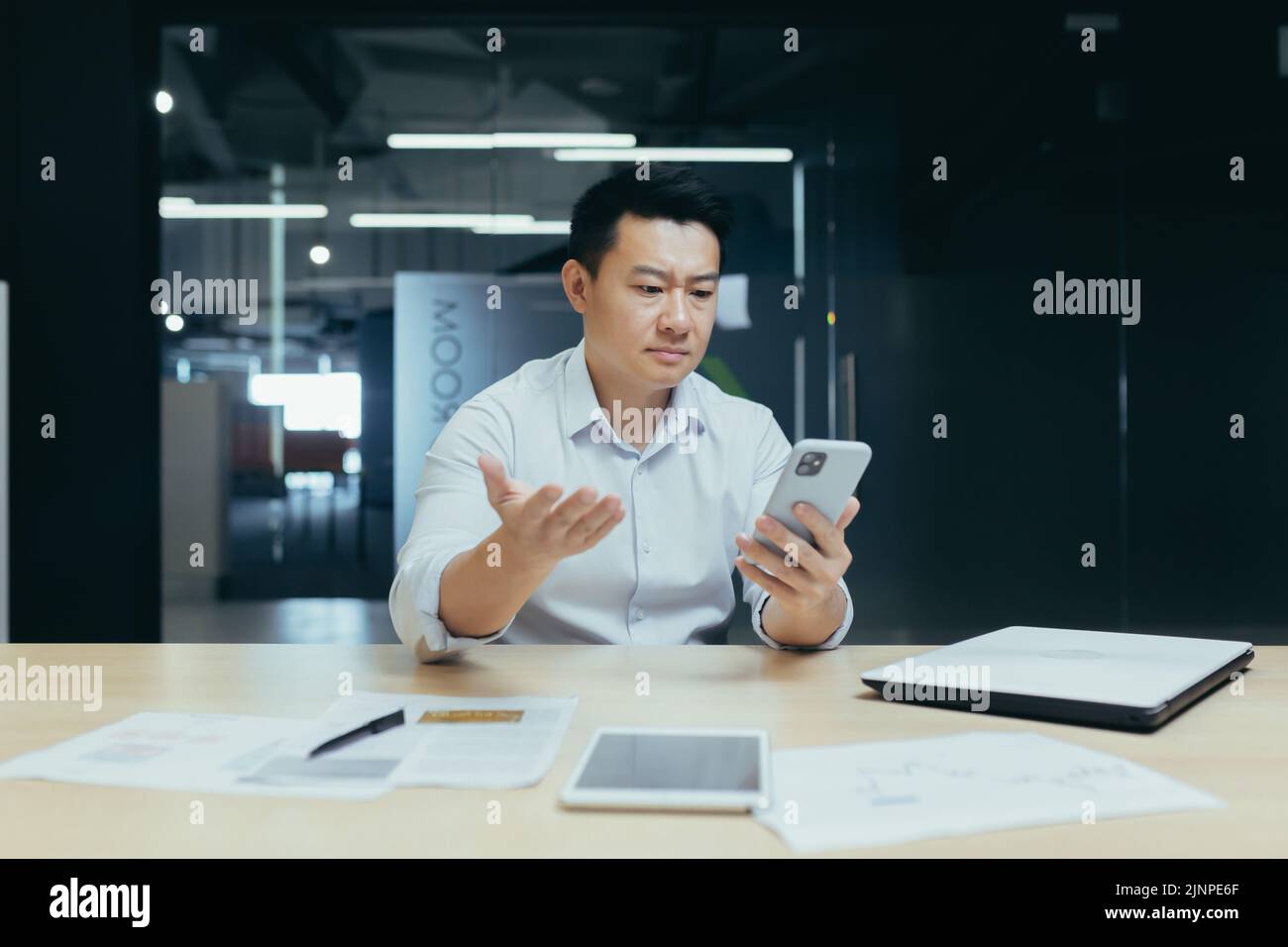 Un ragazzo asiatico confuso che tiene in mano uno smartphone si sente preoccupato di pensare al messaggio ricevuto. Utilizzo quotidiano del telefono cellulare, notizie spiacevoli, attesa di una chiamata importante, concetto di problemi di dispositivo con segnale basso Foto Stock