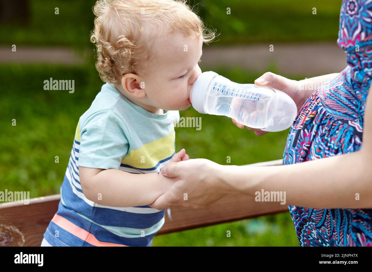 La madre nutre il suo bambino dalla bottiglia con acqua nel parco cittadino. La cura della madre per il bambino. Messa a fuoco selettiva, sfondo sfocato Foto Stock