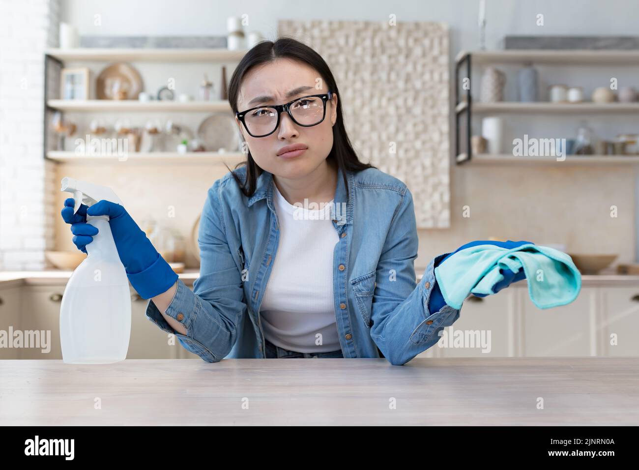 Ritratto di insoddisfatto giovane donna asiatica, casalinga in occhiali guardando la macchina fotografica tenendo detergente e spugna Foto Stock