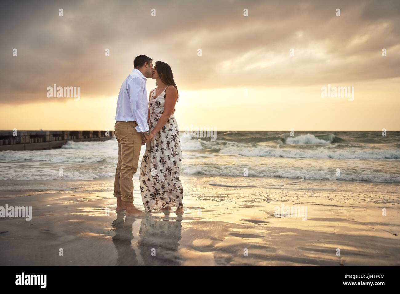 Bacia al tramonto. Scatto completo di una giovane coppia affettuosa che bacia mentre si trova in piedi sulla spiaggia. Foto Stock