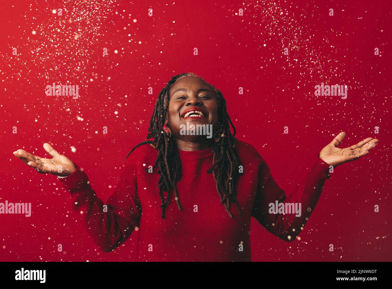 Donna gioiosa con greggi sorridenti eccitatamente mentre si trova sotto i confetti che cadono. Donna matura felice che celebra la vita mentre si trova contro un rosso Foto Stock