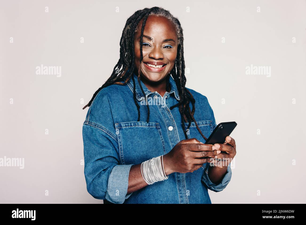 Donna matura che sorride alla fotocamera mentre tiene uno smartphone e si trova su uno sfondo grigio. Donna felice con i Dreadlocks che portano un jack di denim Foto Stock
