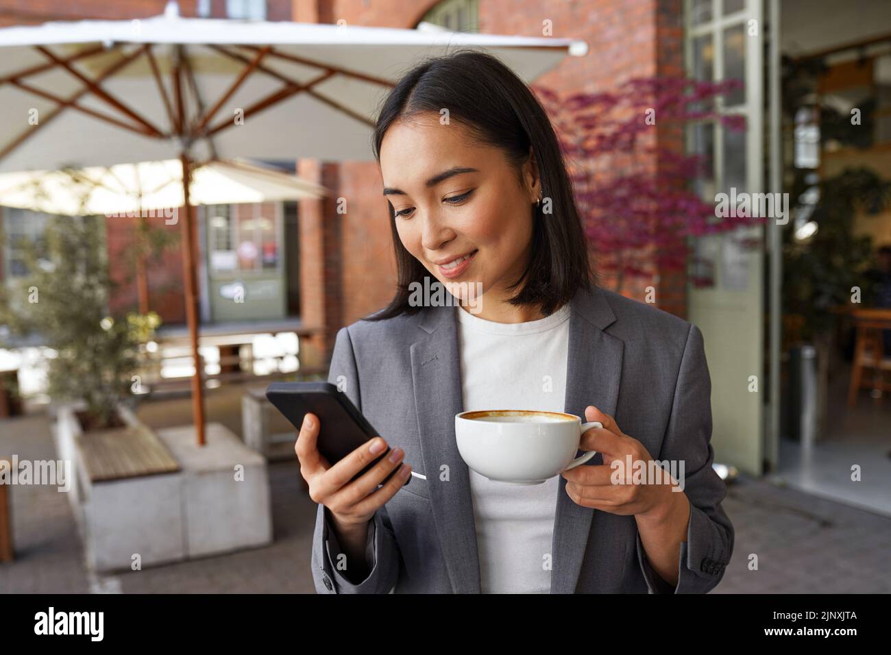 Giovane ed elegante donna asiatica che tiene il caffè in piedi all'aperto utilizzando lo smartphone. Foto Stock