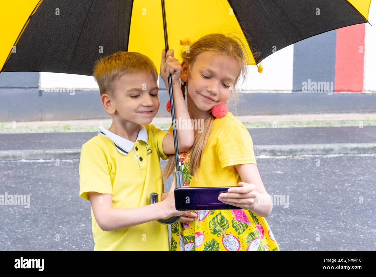 Ritratto di due bambini sorridenti, un ragazzo e una ragazza che tiene un telefono, uno smartphone in piedi sotto un ombrello in un parco in estate. I bambini giocano un onli Foto Stock