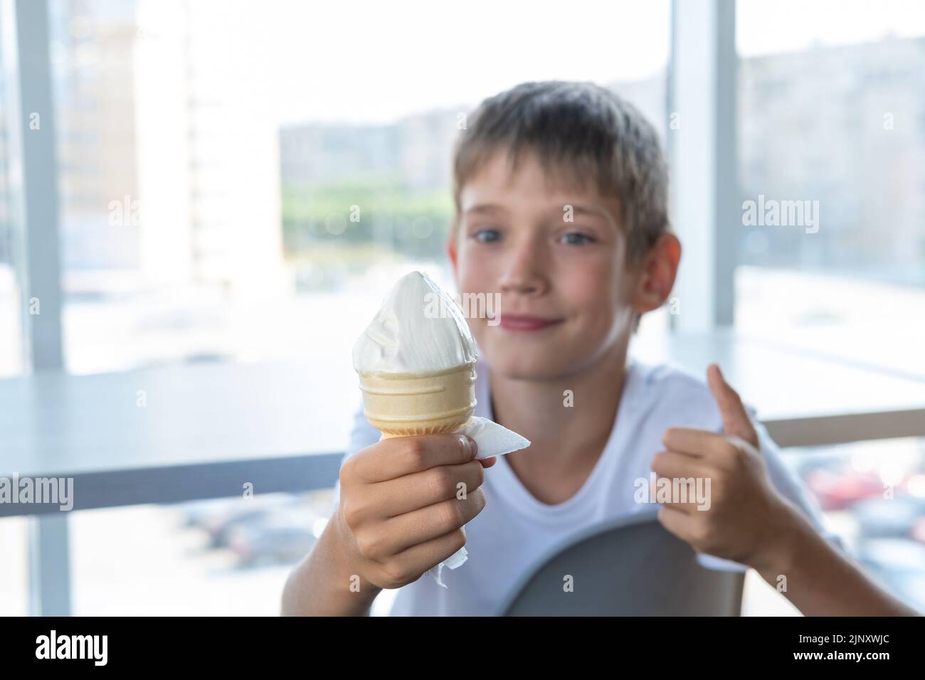 Un ragazzo carino mangia gelato bianco in una tazza di waffle e mostra un gesto di pollice-su mentre si siede a un tavolo vicino alla finestra in un caffè. Sfondo sfocato. A. Foto Stock