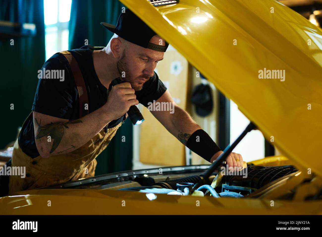 Giovane meccanico di officina di riparazione auto che controlla il motore di automobile gialla mentre dirige flashligh sul relativo motore Foto Stock