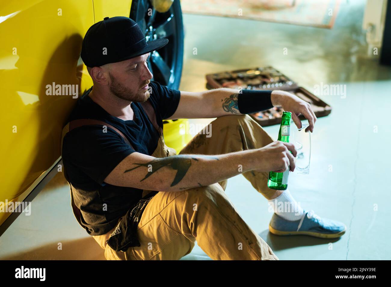 Un giovane riparatore o tecnico stanco con una bottiglia di birra che si riposa sul pavimento del garage mentre si siede in macchina gialla dopo la riparazione Foto Stock