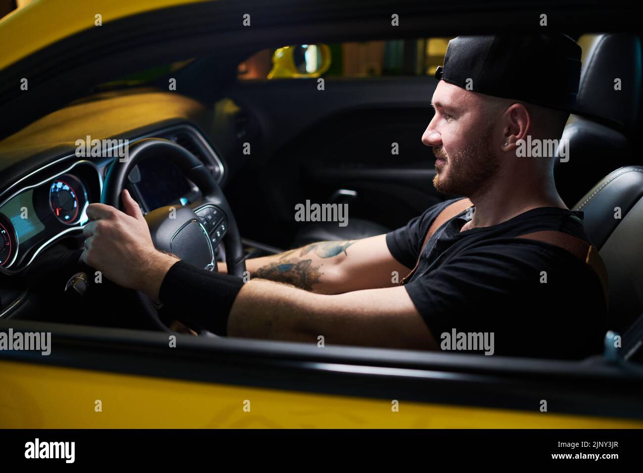 Felice giovane uomo con t-shirt nera e berretto da baseball che tiene il volante mentre si siede sul sedile anteriore dell'auto elettrica durante la prova su strada Foto Stock