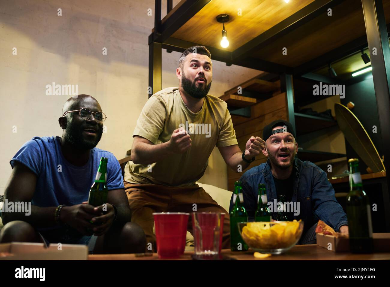 Tre giovani amici multiculturali esprimono la loro gioia per la partita di calcio mentre guardano la trasmissione in tv in garage Foto Stock