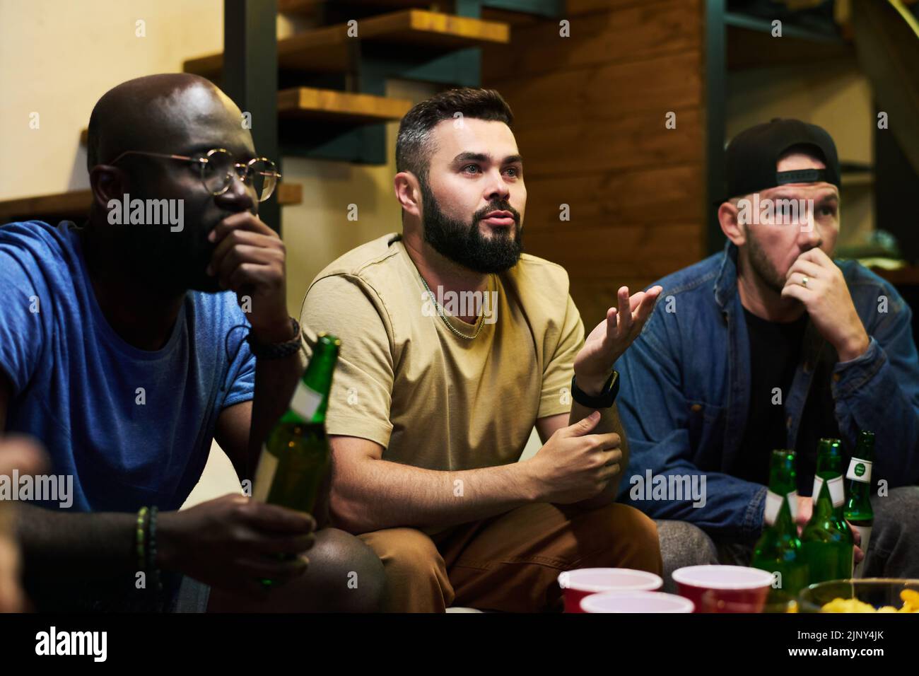 Amici interculturali teso con birra seduto al tavolo e guardare la trasmissione di partita di calcio, mentre uno di loro dire qualcosa Foto Stock