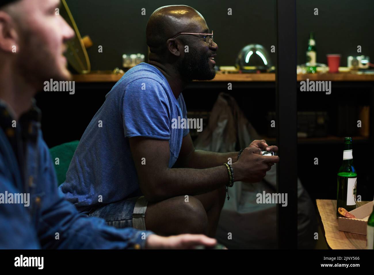 Felice giovane afro-americano uomo in t-shirt blu giocare a video gioco mentre si siede sul divano e premendo i pulsanti sul gamepad Foto Stock