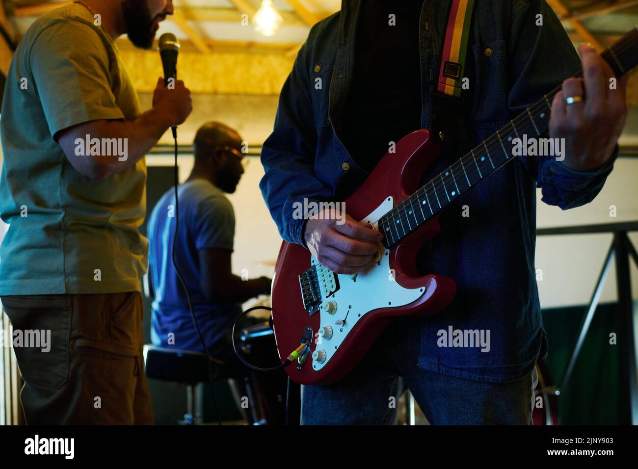 Primo piano di un giovane uomo in giacca denim e jeans blu che suonano chitarra elettrica contro cantante con microfono e batterista Foto Stock